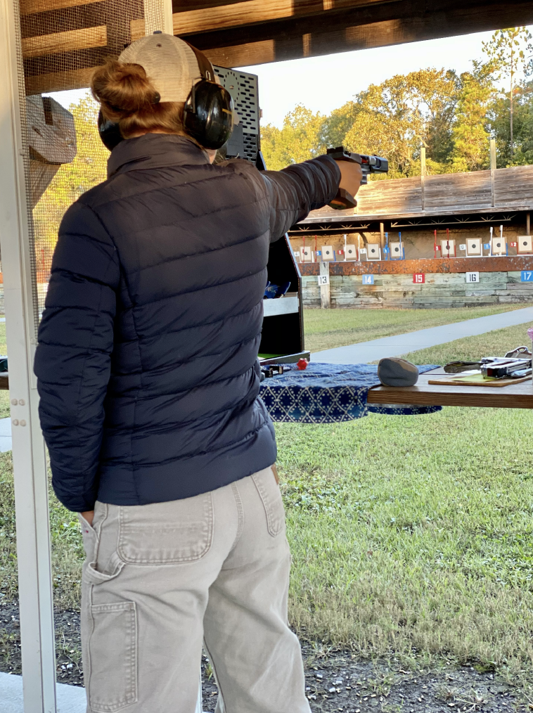 Athlete holding a pistol pointing it at a paper target while competing in a competition wearing a black jacket and brown pants. 