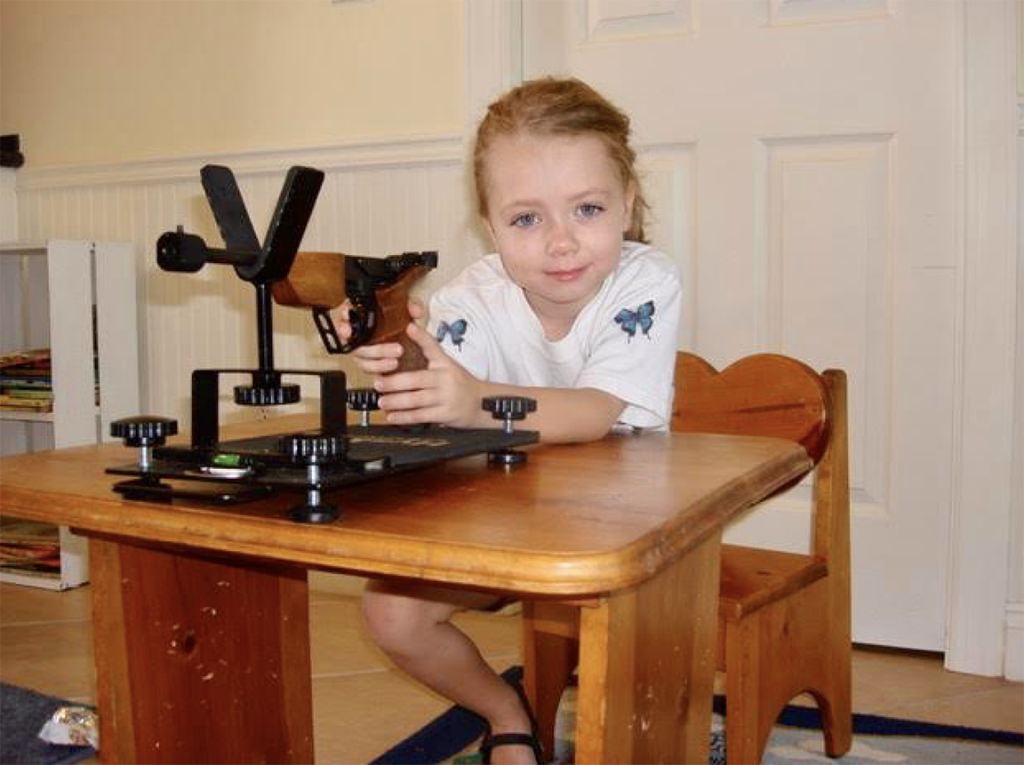 Young girl sitting at a wooden desk in a butterfly shirt while safely holding an air pistol in a brace and smiling. 