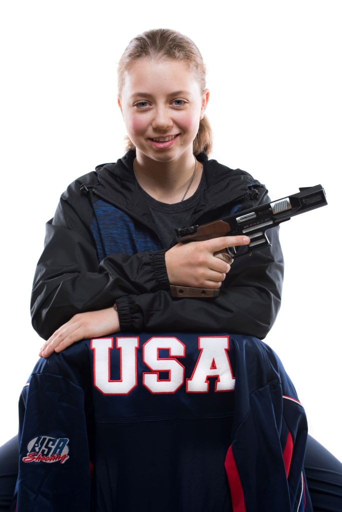 Girl sitting on a chair backwards and holding a safely holding a pistol and the back of her chair has her USA Shooting jacket. 