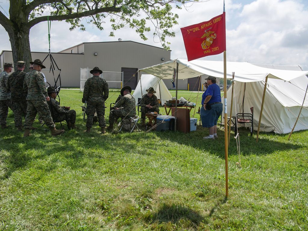 Red guidon flag in front of a tent to let guests know that they are welcome to come hang out with Marines and competitors in the background by the tent and the tree standing. 