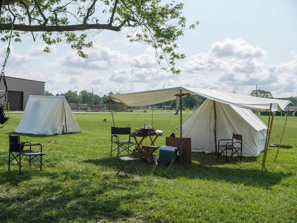 Two tents with chairs out front set up next to a tree in a green field.