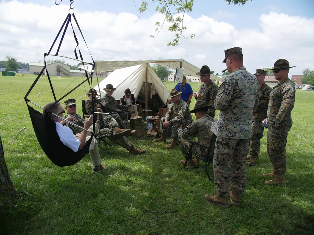 Marines and competitors standing and sitting around outside of a tent and a chair swing in a field on a sunny day.