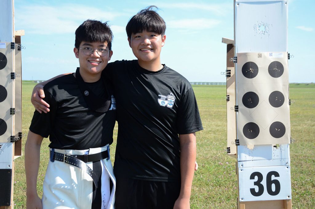 Ryan and Tyler, two teenage boys, stand together outside between paper target stands and smile for the camera.