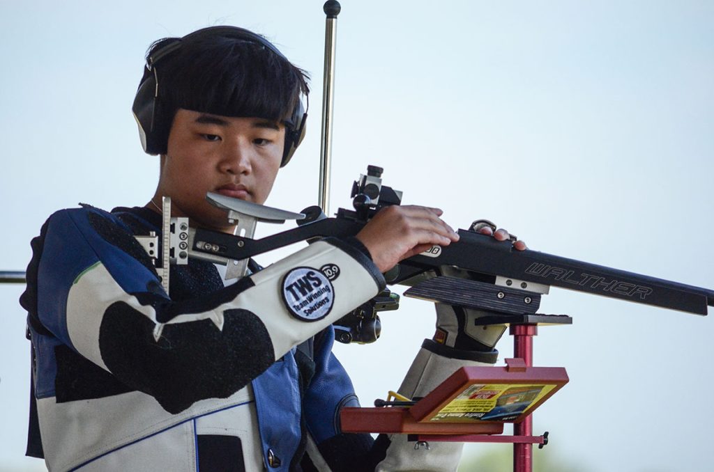 A close up photo of Tyler in the standing position. He has his smallbore rifle resting on his offhand stand and is loading a shot.
