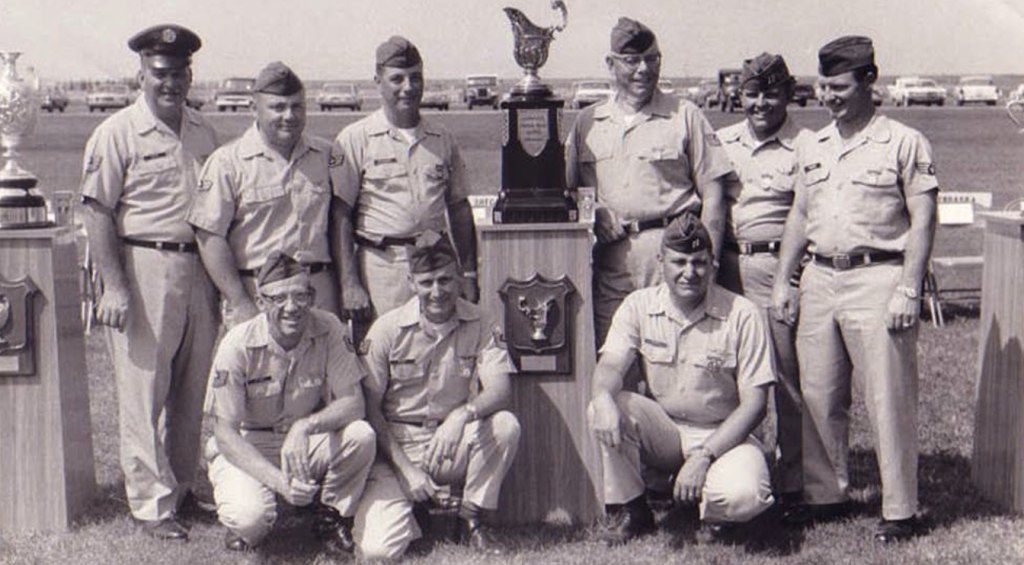 A black and white photograph of the 1966 USAF National Pistol Team. There are nine men in uniform posed together outside with a trophy.