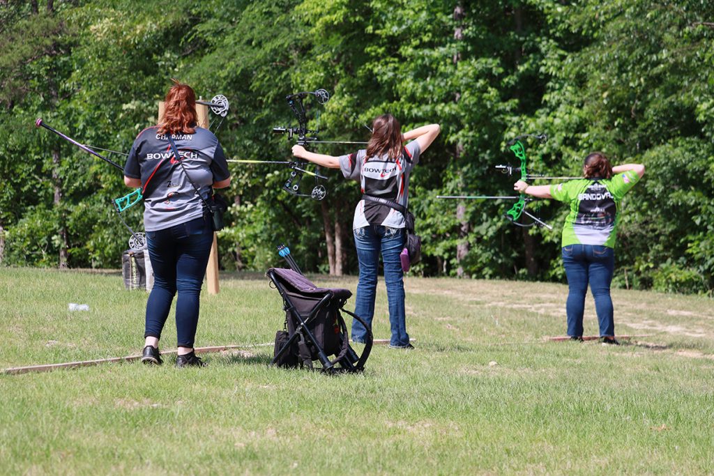 Three women standing on the archery range, two of whom are aiming downrange.