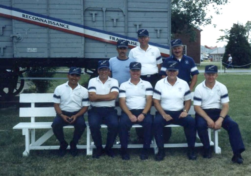 The members of the 1988 United States Air Force National Pistol Team pose outside at Camp Perry. Five men are sitting on a bench and three more men stand behind them.