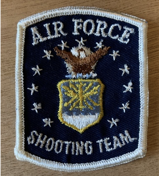 The original United States Air Force Shooting Team logo patch. The patch has a white border and white lettering reading 'Air Force Shooting Team.' A white circle of stars surrounds an eagle over a shield.
