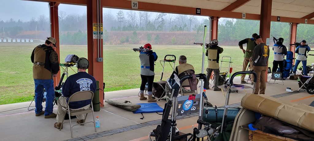 Several competitors standing on the firing line and aiming rifles downrange.