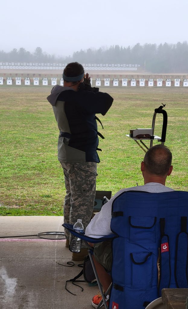 A competitor standing on the firing line, aiming a rifle at electronic targets downrange.