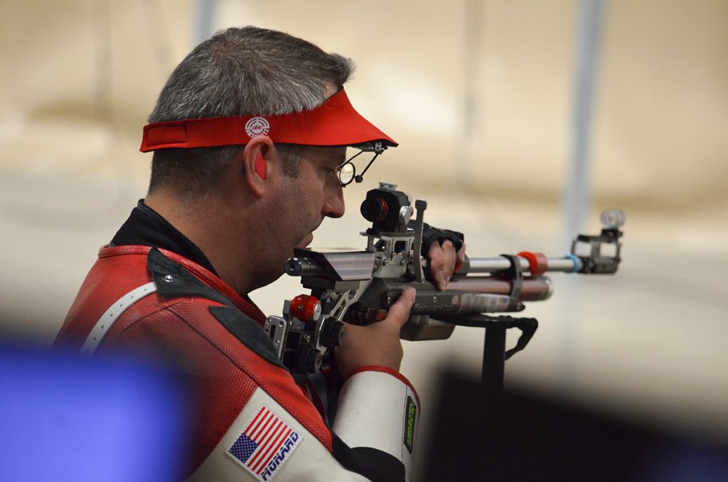 Brian Parziale shoots rifle in a red and white suit.  He is resting his chin against the cheek piece while he centers himself for his air rifle match.