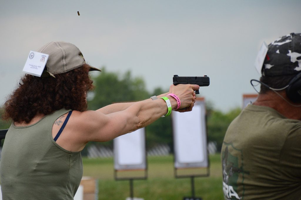 Athlete shoots an outdoor pistol match in a green tank top and brown hat.  Her brass is flying through the air having recently been ejected to chamber the next round and continue firing. 