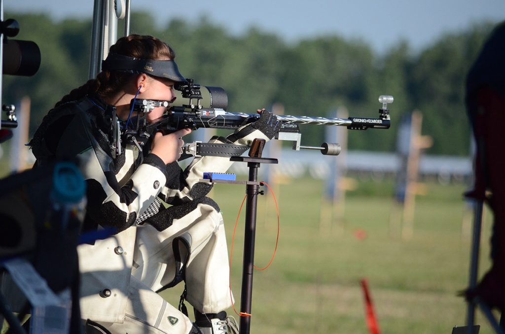 Smallbore shooter in a white and black suit fires from the kneeling position as the sun rises over a green Camp Perry range. 