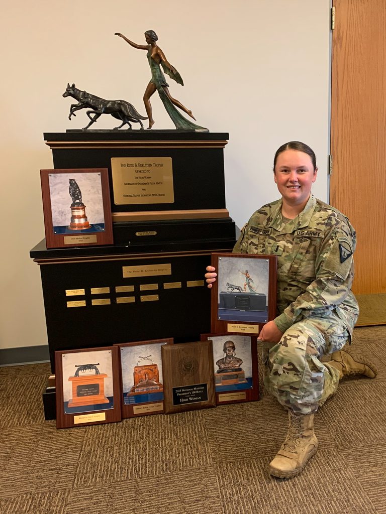 1st Lt. Lisa Emmert Traciak with multiple award plaques posing by the Rose Krelstein Trophy.