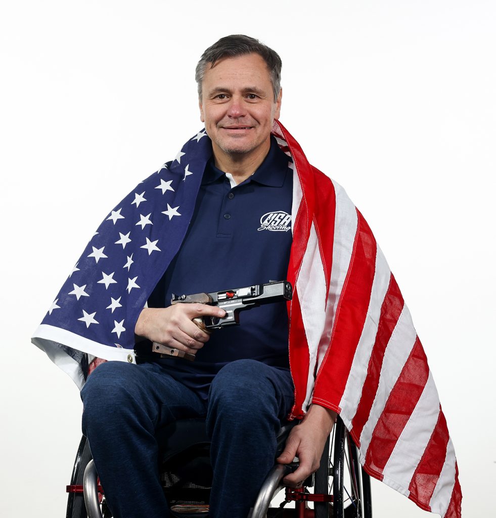 Michael Tagliapietra with the American flag draped over his shoulders, holding his pistol.